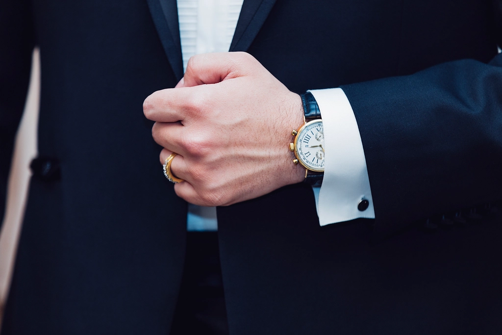 A man in a navy suit fastening his shirt cuff, wearing a watch